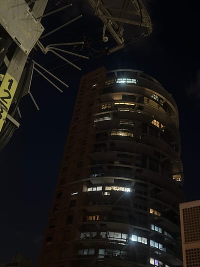 The image presents an upward view at night, featuring two main subjects: a curved residential building and industrial equipment. The building, appearing to be in Givatayim, Israel, has a distinctive curved facade with numerous windows, some illuminated from within. The industrial equipment, located to the left, consists of metal structures with protruding poles. The environment is dark, suggesting an outdoor setting at night. No people are directly visible in the frame.