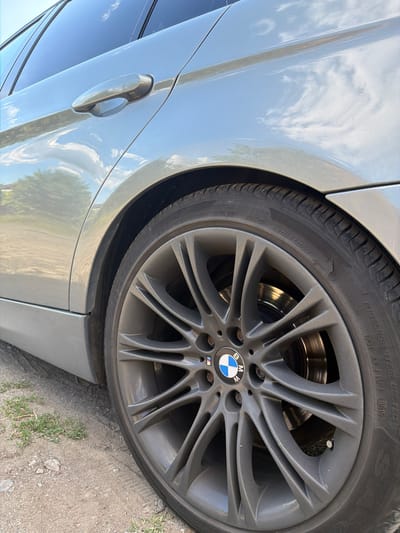 An external, low-angle, close-up, eye-level shot captures the rear passenger-side wheel and lower body panel of a silver BMW sedan. The car is parked on a dirt and gravel path with sparse patches of green grass, likely on the outskirts of Rusokastro, Bulgaria, given the environmental context. The bright midday sun reflects off the car's metallic paint and the polished brake rotors, creating a slightly hazy, bright atmosphere. The tires are black with white lettering, and one of the tires exhibits a small crack. The car's dark tinted windows show reflections of the sky and trees. No people or actions are visible in the image.