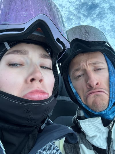 A close-up photograph taken in Saint-Martin-de-Belleville, France, depicts two individuals, one female and one male, positioned side-by-side, likely in a selfie orientation. Both subjects wear black ski helmets with their goggles pushed upwards onto the helmets. The female's goggles, positioned on the left, are purple with a visible texture and show numerous water droplets. The male's goggles, on the right, are dark-tinted and also exhibit water droplets.
The female has fair skin, pronounced eyelashes, and her lips are pursed in a pout, with her eyes partially squinted. She wears a black neck gaiter with perforated sections covering her chin and lower face, and dark winter outerwear. The male has fair skin, facial stubble, and his lips are also pursed in a pouting or grimacing expression, with his eyes partially squinted. He wears a blue patterned balaclava underneath his helmet and a black and white plaid zippered jacket over a white ribbed garment. The background is out of focus, displaying a light, possibly overcast sky or snowy environment, consistent with precipitation.