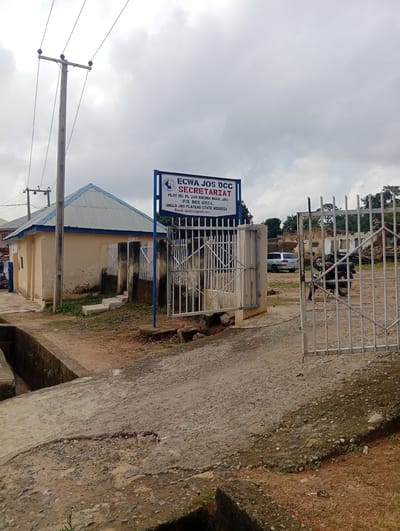 The image shows the entrance to the ECWA JOS DCC Secretariat in Kabong, Nigeria. A weathered beige building with a corrugated metal roof is visible on the left. A metal gate, partially open, leads into a courtyard where a silver car and a motorcycle are parked. A signpost near the gate reads "ECWA JOS DCC SECRETARIAT" with an address and P.O. Box number. The ground is a mix of dirt, gravel, and cracked concrete. Overhead, power lines extend across a cloudy sky. The scene suggests a functional but somewhat unkempt administrative complex.
