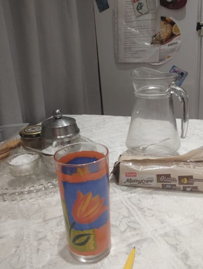 The image shows a table covered with a white lace tablecloth in Nova Dofinivka, Ukraine. On the table, there is a clear glass pitcher with a handle, containing a spoon. To the left of the pitcher, there is a glass with a painted design of a tulip, and a small glass bowl filled with white crystals, possibly salt. Behind these items, a silver sugar bowl with a lid and a glass jar are visible. In the foreground, a package of cookies with the brand name "Yarych" is placed. The background features a gray curtain and a white refrigerator with several magnets and papers attached. The lighting is dim, suggesting an indoor setting, possibly a kitchen or dining area.