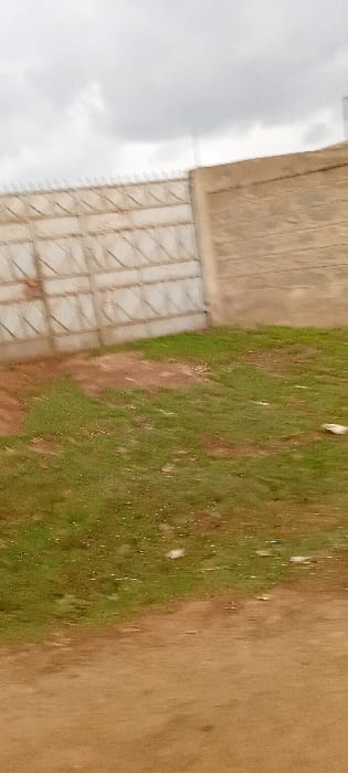 The scene depicts an outdoor environment in Kinale ward, Kenya, under an overcast sky. In the upper portion, a light-colored metal gate with horizontal and diagonal "X" patterns spans the left-center, topped with an array of sharp anti-climb spikes. To its right, a light brown textured concrete or masonry wall abuts the gate. The ground immediately in front of the gate and wall consists of a sloped area featuring patches of green grass interspersed with reddish-brown soil. The foreground is dominated by a flat, reddish-brown dirt or gravel surface, likely a path or road. No individuals, actions, or specific interactions are visible.