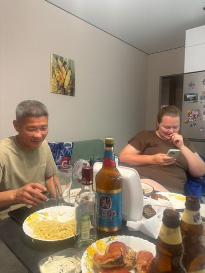 The image depicts an indoor scene, likely a home dining area, with two individuals seated at a table. A man with short, grey hair, wearing a light brown t-shirt, is seated on the left, looking down and holding a glass. To his right, a woman with light brown hair tied back, wearing a dark brown, ribbed t-shirt, is seated and looking at her phone. The table is laden with food and drinks, including plates of spaghetti, roasted food items, bottles of alcohol, and glasses. In the background, a refrigerator is visible with various magnets and pictures attached to its door. A painting of tropical leaves hangs on the wall. The overall impression is of a casual meal or gathering. The location is identified as Novorossiysk, Russia.