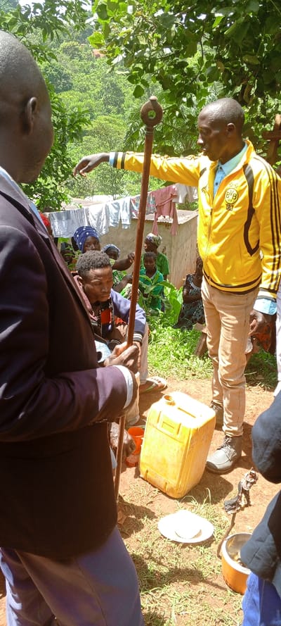 An outdoor gathering takes place in Kundu 1, Democratic Republic of the Congo, under bright sunlight and lush green foliage. In the foreground, partially visible from the back, is a man wearing a dark suit jacket, light blue shirt, and light blue trousers. He holds a long, brown wooden staff with a distinctive circular top. To his right, facing left, stands another man in a vibrant yellow jacket with black stripes (bearing "BVB 09" and "1&1" logos, indicating a Borussia Dortmund football club jacket), a light blue collared shirt, and light brown trousers. He is actively gesturing or pointing with his right hand towards an unseen area to the left.

Seated below him and to the left, a younger man with dark, braided hair wears a dark blue/grey hooded top and looks downwards. Behind these central figures, a group of women and children are gathered, some partially obscured by vegetation. One woman in a blue head covering looks directly at the camera. In the mid-ground, a clothesline with various items of laundry hangs between simple, light-colored structures with corrugated iron roofs.

On the dusty ground, near the feet of the standing man, rests a yellow plastic jerrycan, open at the top. Beside it are two white plates and an orange pot or container holding dark contents. The scene suggests a community meeting or traditional gathering, with the staffs possibly indicating a ceremonial or authoritative role for some participants. The natural environment is dominated by dense trees and greenery, hinting at a rural or forested location.