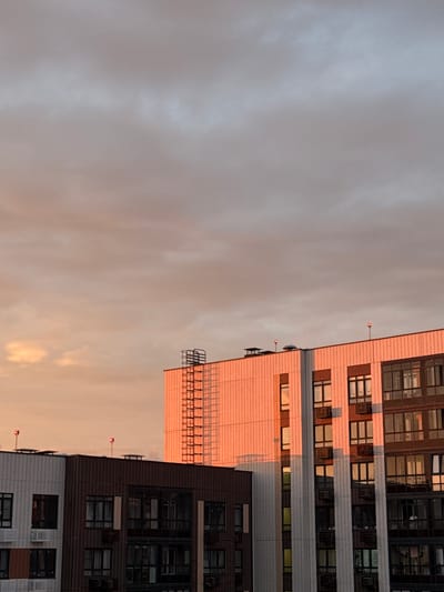 This media file depicts a low-angle view of modern residential buildings in Lyubertsy, Russia, during either sunrise or sunset. The buildings are tall, with multiple stories characterized by numerous windows. The exterior of one building appears to be clad in light-colored tiles, reflecting the warm hues of the sky, while another section features dark brickwork. Architectural details include balconies, external staircases, and ventilation units. The sky is overcast with soft, diffused light, suggesting a cloudy day or the transition of light during dawn or dusk. The overall mood is serene, with the warm light cast upon the buildings creating a peaceful atmosphere. There is no visible text within the frame.
