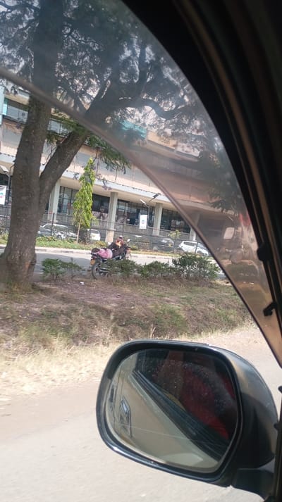 The image depicts an outdoor urban scene in Nakuru, Kenya, viewed from inside a vehicle through its windshield. The passenger-side mirror, showing a crack and reflecting a red interior, is visible in the lower right quadrant. A paved road traverses the mid-ground. To the left of the road, a large tree with a thick trunk stands on a grassy embankment. Further along the embankment, a person dressed in dark clothing is seated on a black motorcycle, which has a pink object secured to its rear. In the background, a multi-story commercial building, identified by a visible sign reading "lladin" (likely "Aladdin"), features a light-colored facade and numerous dark-paned windows. Multiple cars are parked in front of this building. The scene is illuminated by natural daylight. Based on the "Aladdin" sign and building architecture, the location is identified as being in front of the Aladdin Golden Palace Hotel in Nakuru.