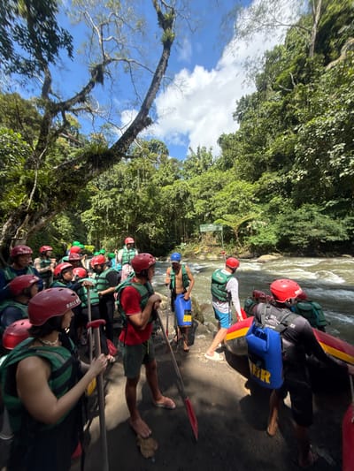 A long line of people in red helmets and striped green and black life vests are waiting to board rafts near a river. Paddles are visible, held by many of the individuals. The rafts are red and black, with blue containers. The scene is in a lush, green environment, consistent with a tropical setting. The river appears fast-flowing. A sign is visible in the background. A tree with aerial roots and a bright blue sky with clouds are on the left side of the frame. The location is Payangan, Indonesia.