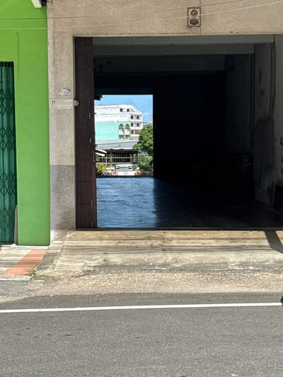 The image shows a narrow street view in Ranong, Thailand. In the foreground, a paved road with a white line runs horizontally. Beyond the road, there is a raised concrete sidewalk with a textured surface. To the left, a bright green building with a metal security gate is visible. To the right of the green building is a light-colored concrete pillar.
The central part of the image features a dark, open entryway or archway. Through the archway, a glimpse of a building with a light-colored, tiered facade and a green roof is visible, with trees and a blue sky in the background. The floor inside the archway appears to be a dark, reflective surface, possibly wet. Wooden planks are visible on the ground leading up to the archway.
There are no people visible in the image. The scene appears to be a commercial or residential street in the city of Ranong.