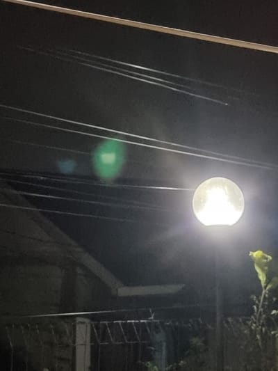 This image depicts an outdoor night scene in Pacet, Indonesia. The dominant feature is a bright, spherical street lamp mounted on a pole, situated on the right side of the frame and emitting white light. Above and to the left of the lamp, multiple dark, parallel utility lines span horizontally across the upper two-thirds of the image. A light-colored, sloping architectural element, possibly a roofline or eave, is visible in the mid-ground, below the utility lines and behind the streetlamp. Below this structure, a dark, patterned metal fence extends horizontally. Sparse vegetation, including leaves, is partially visible on the far right, illuminated by the streetlamp. Two prominent lens flares, one green and one smaller blue, are present in the upper-left quadrant of the image. No individuals or active subjects are present.
