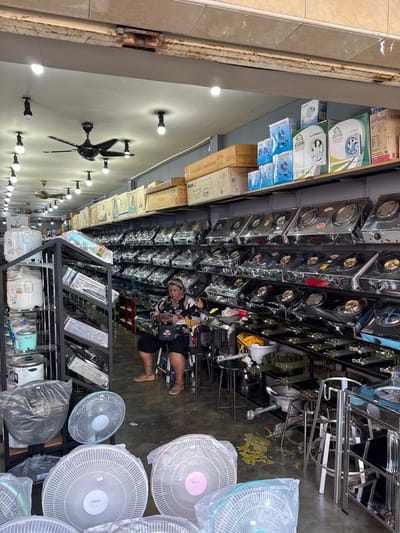 The image shows the interior of a retail store in Ranong, Thailand, specializing in kitchen appliances. The store is brightly lit with overhead fluorescent lights and ceiling fans. Rows of gas stoves are displayed on shelves, most covered in plastic wrapping. Various kitchen appliances, including rice cookers and electric fans, are also visible throughout the store. A woman is seated on a stool near the shelves of stoves, looking at her phone. The floor is concrete and appears to have some yellow staining. The overall environment suggests a busy marketplace or commercial district within the city.