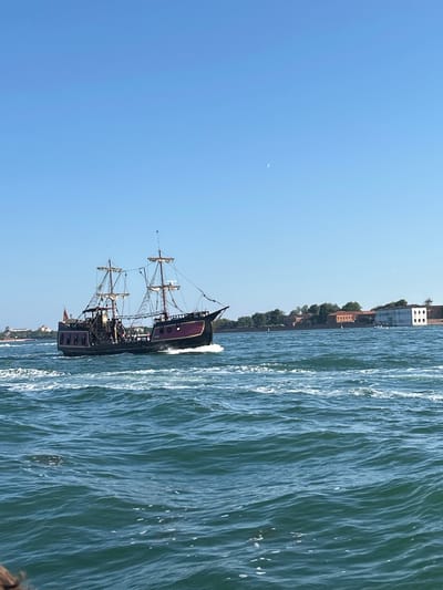 This image depicts a scenic view of a tall ship sailing on a body of water, likely a canal or bay, with a cityscape in the background. The sky is a clear, bright blue, suggesting a sunny day with good weather. The water is a deep blue-green, with noticeable ripples and whitecaps indicating movement. The tall ship is the primary subject, featuring multiple masts and sails, and appears to be an older, perhaps historical, vessel, potentially designed to resemble a pirate ship. It has dark colored hull with some ornate details and windows. The ship is in motion, creating a wake behind it.
In the background, a line of buildings and trees can be seen along the shore. The architecture suggests an urban environment, and the mention of Venice as a location context strongly implies the setting is within the city's famous waterways. The buildings are varied in style, some appearing older with brickwork, while others are more modern, painted white. Green trees are interspersed among the structures.
The time of day appears to be midday or afternoon, given the bright sunlight and clear sky. There are no visible people in the immediate foreground, nor is there any text present in the image. The overall impression is one of a picturesque day in a historic Italian city, with a striking nautical element dominating the scene. The image's orientation is portrait, with the ship and water filling the right side of the frame, and the sky and distant land on the left.