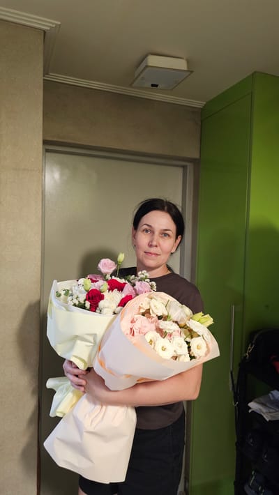 A woman of East Asian descent is pictured holding two bouquets of flowers. She has dark hair and is wearing a dark t-shirt and black pants. She is smiling and looking towards the camera. The bouquets are large and colorful, with a variety of flowers including roses, lisianthus, and chrysanthemums. The flowers are wrapped in pale pink and yellow paper. The background appears to be indoors, with a green cabinet and a door visible. There are also some bags and clothing items on top of the cabinet. The lighting suggests it is daytime. The overall mood of the image is happy and celebratory. The woman is standing, and the image is taken from a slightly low angle.