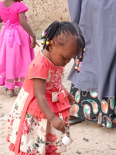 This photograph captures a candid outdoor scene featuring several individuals, likely in Rangaza, Nigeria. The central subject is a young Black girl, possibly a toddler, dressed in a colorful and elaborate outfit. She wears a coral red top with ruffled layers and delicate floral embroidery, paired with a cream-colored skirt adorned with a prominent red and green floral pattern. Her hair is styled in neat braids, decorated with numerous brightly colored beads in shades of yellow, pink, green, and blue, which also dangle from the sides of her head. She is looking downwards with a focused expression, holding a small white scoop or spoon in her right hand.
To her left, partially visible, is another young girl in a vibrant, floor-length hot pink dress with a large bow tied at the back. Her activity is unclear, but she appears to be looking down or to the side, with her right hand slightly extended. On the right side of the frame, the lower portion of an adult figure is visible, wearing a flowing grey garment, possibly an abaya, over a skirt with a distinctive black background featuring large, colorful paisley-like patterns in shades of orange, teal, and cream.
The setting is an outdoor space with light-colored, uneven ground, suggesting dirt or sand. A textured, light-colored wall with rough, irregular patches forms the background. The natural, bright lighting indicates it is daytime. There is no visible text in the image. The scene conveys a sense of daily life and children engaged in simple activities within their environment.