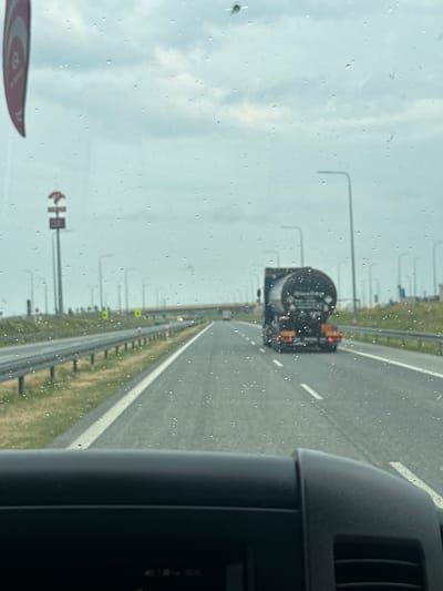 The image shows a view from inside a vehicle looking out through a rain-streaked windshield at a highway. A dark-colored tanker truck is traveling ahead on the right lane of the highway. The sky is overcast. On the left side of the road, there are streetlights and a signpost with a circular logo and a stylized “H”. The scene appears to be on a highway in or near Chomentowo, Poland, based on visual cues and context. The dashboard of the vehicle is visible in the foreground.
