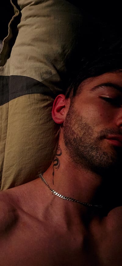 The image shows a close-up, overhead view of a person lying down, possibly sleeping. The person has dark hair and stubble on their face. A tattoo of a snake is visible on their neck, and they are wearing a silver chain. The background appears to be a pillow or bedding with a patterned texture. The lighting is dim, with shadows creating a dramatic effect. The scene is likely indoors, given the presence of bedding. No specific location in Kostenets, Bulgaria is depicted or identifiable from the image.