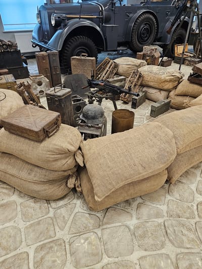 The image presents a collection of military artifacts and equipment arranged against a stone-tiled floor, possibly an exhibit or museum display. Dominating the scene are several burlap sacks, stacked and bundled together. Adjacent to these, there's an array of military-related objects including ammunition crates, metal containers (possibly fuel canisters), a soldier's helmet, and what appears to be a machine gun mounted on a stand. A vintage military vehicle, possibly a truck, is partially visible on the left side of the image, showcasing its tires and front section. The setting is likely an indoor environment. This image shows a display at the Vadim Zadorozhny Technology Museum in Архангельское, Russia.