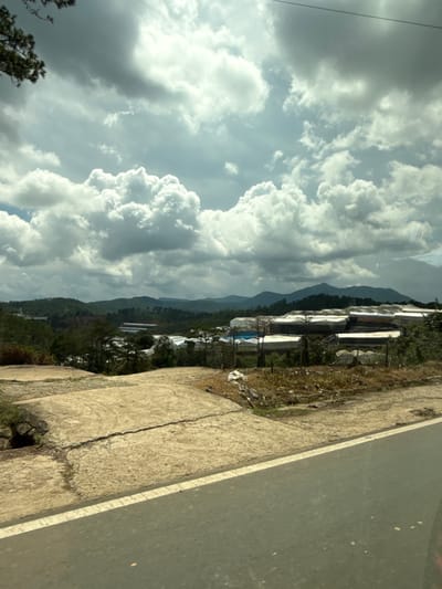 The image depicts a landscape scene in Thôn 6, Vietnam, under a dramatic sky filled with both dark, heavy clouds and bright, sunlit patches. In the foreground, a paved road with a white line runs diagonally from bottom right to top left. Adjacent to the road is a rough, uneven, light-colored ground surface with cracks and vegetation.
Further into the scene, a vast area of greenhouses with white, translucent roofs is visible, stretching across the middle ground. These structures are arranged in rows and appear to be part of an agricultural or industrial complex. Behind the greenhouses, rolling green hills and mountains form the backdrop, with more greenery and trees dotting the landscape. The overall impression is one of a rural or semi-rural area experiencing dynamic weather conditions. No people are discernible in the image.