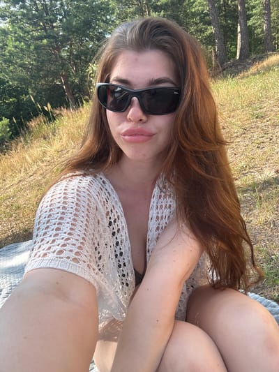 A young woman with long, reddish-brown hair and light skin is sitting on a blanket outdoors. She is wearing black sunglasses and a white crocheted top over a dark bikini. The background shows a grassy slope with trees, suggesting a park or natural area. The lighting indicates it is daytime, likely sunny. The location is identified as Kurovskoye, Russia.