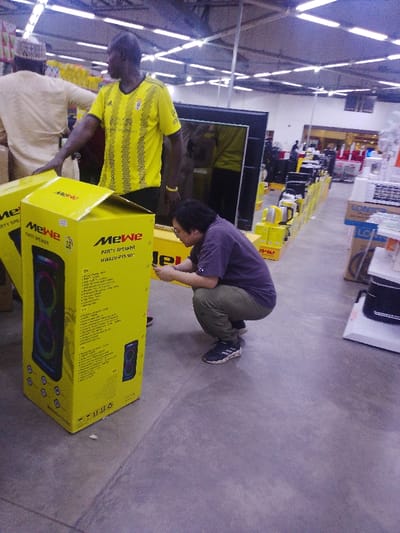 The image captures an indoor scene within an electronics retail store in Gandu Albasa, Nigeria. Three individuals are prominently featured. In the foreground, a person of East Asian descent, wearing a dark purple short-sleeved shirt, olive green cargo pants, and black and white athletic shoes, is in a full squat position, focused on a smartphone held in both hands. To their left, an adult male of African descent stands upright, dressed in a yellow and black striped short-sleeved athletic shirt with a white emblem and a yellow wristband. He gazes towards the right of the frame, with his left hand resting near two large, bright yellow cardboard boxes. These boxes are labeled "MeWe PARTY SPEAKER MWAUD-PTS100"; one is partially open, revealing a dark speaker unit with multi-colored lights. In the background, partially obscured, an adult male of African descent wearing a light beige traditional Nigerian garment and cap is seen facing away, appearing to interact with merchandise on a display. The store environment includes a plain concrete floor, a ceiling with visible ductwork and evenly spaced fluorescent strip lighting, and multiple rows of electronics, including large flat-screen televisions and smaller appliances displayed on yellow stands and shelves, extending into the background.