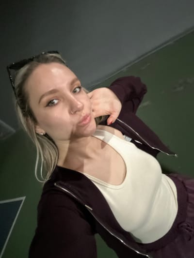 A woman is seen in a selfie, wearing a beige top and a zipped purple jacket. She has blonde hair and is wearing sunglasses on top of her head. The background is a green surface with a gray wall on the left. There is no indication of a location in Esenyurt, Turkey.