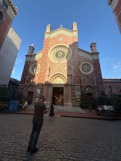 An eye-level, wide shot captures the facade of the Basilica of Saint Anthony of Padua in Beyoğlu, Turkey, bathed in sunlight under a clear blue sky. The large brick church features a central rose window and smaller circular windows with intricate stonework. Flanking the main entrance are arched doorways with mosaics above. Several potted plants adorn the area in front of the church, and a large informational sign is visible to the left.

In the foreground, a man with short hair and glasses stands with his back to the camera, wearing a dark olive t-shirt and dark jeans. He appears to be holding a phone. To his right, a woman in a dark patterned dress walks towards the church entrance. Further to the left, two women are seen near the informational sign. A statue of a robed figure stands to the right of the church entrance, partially obscured by foliage. The ground is paved with cobblestones.
