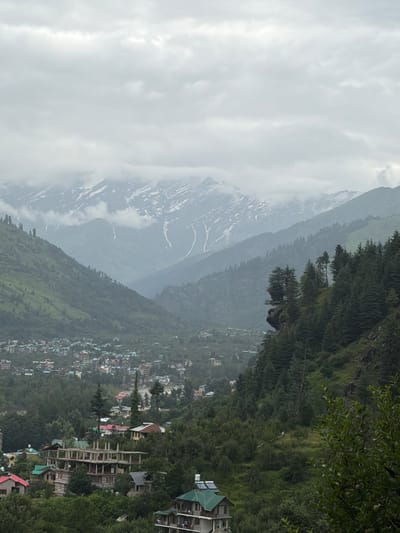 The image depicts a panoramic view of the city of Bahang, India, nestled within a valley surrounded by lush green hills and towering snow-capped mountains. The sky is overcast with heavy clouds, casting a soft, diffused light over the scene. In the foreground, a cluster of buildings, some appearing to be under construction, dot the hillside, interspersed with dense foliage and tall pine trees. The town of Bahang is visible further down in the valley, characterized by a dense arrangement of houses with colorful roofs, suggesting a vibrant community. The distant mountains form a majestic backdrop, with patches of snow visible on their upper slopes, indicative of a high-altitude location. The overall impression is one of a serene and picturesque landscape, where human settlement harmoniously coexists with the grandeur of nature.