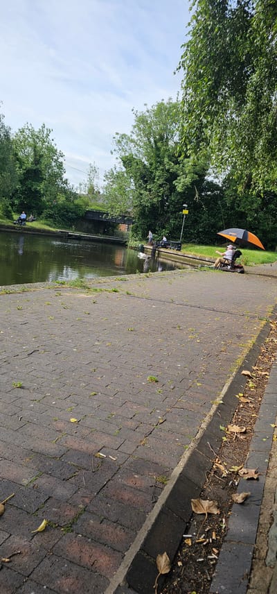 The image captures a scene at the Montgomery Canal in Welshpool, United Kingdom. In the foreground, a brick-paved walkway borders the canal. Scattered across the pavement are small weeds and fallen leaves. The canal itself is visible, with its dark water reflecting the overcast sky. Along the canal bank, on the left, a person sits in a mobility scooter amidst green grass. Further along the canal, a pedestrian bridge with a metal structure spans the water. On the right side of the canal, several individuals are present. One person is seated in a chair under a large orange and black umbrella, holding a fishing rod. Another person is fishing nearby, also seated. The background is dominated by lush green trees and vegetation lining the canal. The sky is a pale blue with scattered clouds.