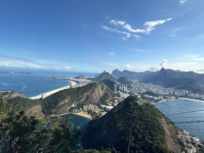 This panoramic high-angle photograph captures a breathtaking view of a vibrant coastal city, likely Rio de Janeiro, nestled between towering green mountains and the expansive blue ocean under a clear sky.
In the foreground, the steep, densely vegetated slope of a mountain dominates the right side, with several parallel cable car lines stretching diagonally across the frame, hinting at an aerial tramway system. Below, a small bay with a sandy beach and some buildings is visible.
The midground unfolds a dramatic landscape. To the left, a long, curving white sand beach, lined with numerous high-rise buildings, meets the deep blue sea. The ocean extends to the horizon, dotted with a few small islands. In the center, another prominent, rocky, tree-covered mountain rises, with urban development clustered around its base. To the right, a large, sheltered bay (likely Botafogo Bay) is filled with hundreds of small boats and yachts anchored in its calm waters, bordered by a bustling cityscape.
The background features a majestic series of rugged, verdant mountains characteristic of the region, towering over the urban sprawl that continues to extend into the distance along the coast and valleys. The sky above is a brilliant azure, adorned with scattered white cumulus clouds, indicating a bright and clear day.
The scene depicts a bustling urban environment harmoniously integrated with stunning natural beauty. No specific human activity is discernible beyond the general presence of a city, boats, and the cable car infrastructure. There is no visible text in the image.