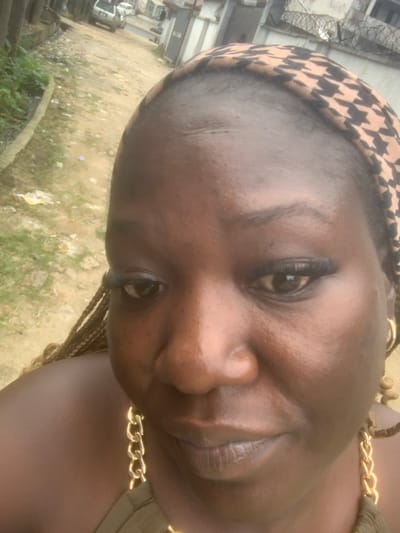 A medium-close-up selfie shows a Black woman with dark skin, braided hair, and a patterned headwrap. She is wearing large hoop earrings and a gold chain necklace. Her makeup includes dark eyeliner and mascara. The background is blurred and appears to be an outdoor urban setting with a dirt road, green vegetation, and buildings. Based on other images from the same source, the location is identified as Eliozu, Nigeria.