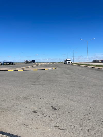 The image shows a view of a parking lot or loading area in Arshaly, Kazakhstan. The area is paved with asphalt and marked with black and yellow barriers. The sky is clear and blue. A semi-trailer truck is parked in the background, along with other similar vehicles in the distance. Streetlights line the area.