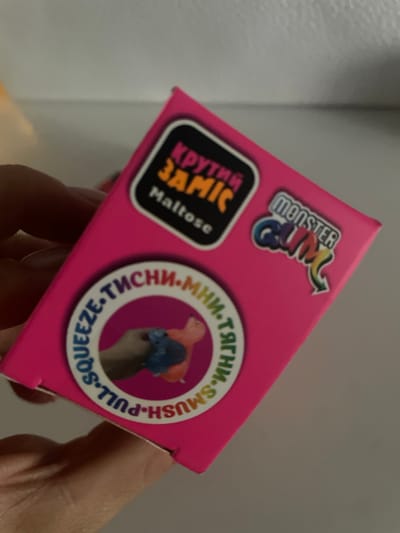 The image shows a close-up of a pink box with various labels and graphics. A person's hand is holding the box, which is angled. On the box, there is a sticker with the text "MONSTER CAM" in colorful letters. Another sticker features the text "КРУТИЙ SAMIC" in large, bold, yellow letters, with smaller text "Maltose" below it. A circular label with text in both Russian and English reads "L-SQUEEZE-ТИСНИ•M HM-SMUSH-PULL-S". Inside this circle, a blurred image shows two small, colorful figures. The background is a plain, light-colored wall. The lighting appears to be artificial, suggesting an indoor setting. The overall impression is that of a product packaging, likely for a toy or craft item, being presented or examined. The presence of Russian text suggests a market that uses this language, while the specific location is noted as Brovary, Ukraine.