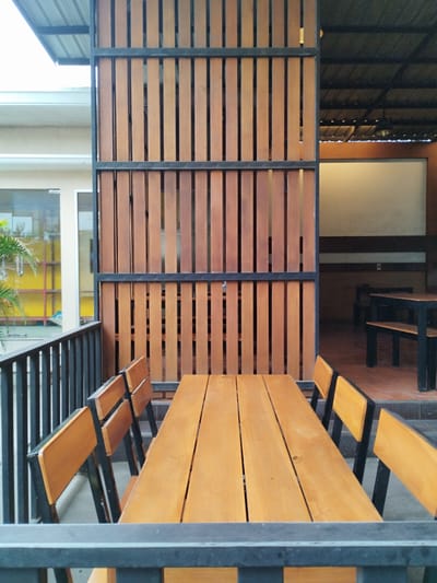 The image shows an outdoor dining area with several wooden tables and benches. The tables are long and made of light-colored wood, with a rustic finish. The benches are also made of wood and have black metal frames. The area is partially covered by a roof, and there is a vertical wooden screen in the background. In the distance, there are more tables and benches, suggesting a larger seating area. The scene appears to be in a well-lit, possibly tropical environment, with greenery visible in the background. The location is identified as Payameuneng, Indonesia.