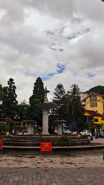 The image shows a fountain in Sa Pa Ward, Vietnam, during an overcast day. The fountain is in the center of a paved area, surrounded by trees and a yellow building labeled "Boutique Hotel." A red sign with white lettering is placed near the fountain, and several cars are parked in the background. People can be seen walking in the distance. The foreground consists of wet cobblestones.