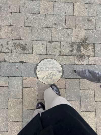 The image shows a close-up, overhead view of a paved sidewalk with a circular bronze plaque embedded in it. The plaque commemorates a horse named Nijinsky, a jockey named Lester Piggott, and their victory in the third leg of the Triple Crown at Doncaster in 1970. The plaque features an engraving of a horse and jockey. The sidewalk is made of rectangular, greyish paving stones with visible grout lines. A person's legs and feet are visible in the lower part of the frame. They are wearing dark-colored dress shoes and light-colored trousers. Partially visible in the upper right quadrant of the image is a folded umbrella with a grey fabric. The overall environment appears to be outdoors, likely in a public area, with the plaque marking a significant historical event related to horse racing, possibly within Epsom and Ewell, known for its racing heritage.