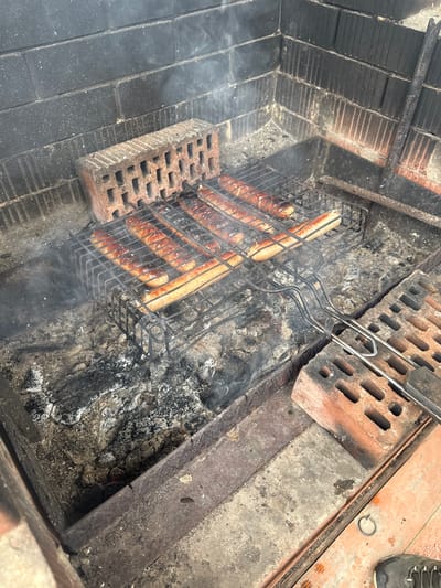In Kurovskoye, Russia, eight sausages are positioned on a black metal grill basket, actively cooking over smoldering charcoal embers and ash within a brick-built outdoor brazier. Smoke rises from the heat source. The interior walls of the brazier are constructed from dark, rectangular bricks, exhibiting soot accumulation. A light-colored, perforated brick supports the left side of the grill basket. On the right, a similar brick supports the metal handle of the grill basket, adjacent to a dark-handled utensil, possibly tongs or a spatula. The ground immediately surrounding the brazier consists of concrete and a reddish-brown surface. The toe of a black shoe with white laces is visible in the lower right foreground.