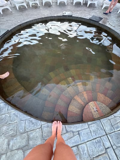 A high-angle shot depicts a round stone pool filled with water. A person is standing next to the pool, their feet and lower legs visible on the right side. A second person stands next to the pool, only their foot visible on the lower left side of the pool. The pool has stone steps leading down into the water. The pool is surrounded by stone pavers. The scene is possibly in Kara-Oy, Kyrgyzstan. A row of white chairs is visible on the left side.