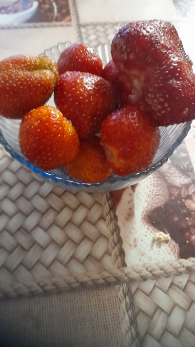 A close-up, eye-level shot shows a glass bowl filled with strawberries. The strawberries are a mix of red and orange hues, with some appearing riper than others. The bowl itself is clear glass with a decorative scalloped edge. The background is a blurred tapestry-like pattern in beige and brown tones, with a rope accent visible in the foreground. No people are present in the image, and there is no indication of the specific location within Bagayevskaya, Russia.