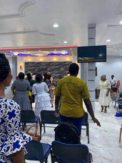 The scene depicts an indoor gathering within an event hall located in Gbawe, Ghana. The interior features a white false ceiling with decorative moldings and recessed circular light fixtures, augmented by red and blue LED strip lighting along a raised section at the front. The prominent front wall is adorned with dark, swirling marble-patterned paneling. The floor is tiled in white with a light gray marble-like pattern.

Approximately 15-20 individuals are present, primarily women, mostly standing. In the foreground left, the partial left side and back of a woman in a blue and white patterned dress and a black head covering are visible. In the foreground center-right, a man wearing a yellow patterned short-sleeved shirt and blue trousers is viewed from the back, holding a small white object in his right hand. A black backpack rests on a dark blue plastic chair directly in front of him.

In the mid-ground, multiple individuals are standing. These include a woman in a dark patterned dress, seen from the back, and another woman in a white tiered ruffled dress, also seen from the back. To the right, a woman in a white lace dress with a yellow floral pattern on the bodice stands with her mouth open, facing towards the right. She is wearing light green shoes.

A flat-screen television is mounted on a gray column on the right side of the room, displaying an image of an open book, possibly a Bible, on a dark background. Dark blue plastic chairs with metal frames are arranged in rows; some are occupied, while others are empty or hold personal items like a brown handbag on a chair in the mid-ground. In the far background, behind the woman in the white lace dress, parts of a red drum and a man in a white shirt are faintly visible.
