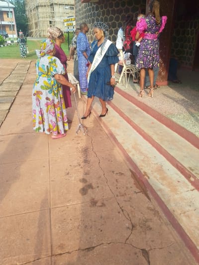 The image shows a group of people outside a building in Awka, Nigeria. A woman in a blue dress and a sash, possibly a beauty pageant winner or titleholder, stands on a step. Next to her, an older woman in a colorful patterned dress uses a quad cane. A few other people are visible, including a woman holding a baby wearing a pink dress, a man in a blue patterned shirt, and other individuals in the background. In the background, a building under construction, scaffolding visible, is situated next to another structure marked "CHAPEL OF THE GOOD SHEPHERD". The ground is paved with tiles, and there are a few cracks in the pavement.