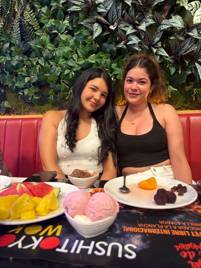 Two women are posing for a photo, seated in a restaurant booth with red leather upholstery. The woman on the left has wavy, reddish-brown hair and is wearing a black tank top and white shorts. She is smiling at the camera. The woman on the right has long, dark hair and is wearing a white top adorned with pearls. She is also smiling.
In front of them on a table are plates of food. One plate contains several small, dark berries and a slice of orange. Another plate holds fruit, including slices of watermelon and pineapple. There are also bowls of ice cream, one appearing to be strawberry flavored and the other chocolate. A menu with Spanish text is partially visible behind the food. The text on the menu includes "BUFFET LIBRE INTERNACIONAL", "RISCADA A LA PLANCHA", "RRILLA ASADOR", and "Sushi Wok".
The setting appears to be indoors, possibly a restaurant with a decorative wall of greenery behind the subjects. The lighting suggests it is daytime, but the exact time is not discernible. The overall mood is casual and happy, with both women appearing relaxed and enjoying their meal. The location context provided indicates the setting is Carretera De San Martín, Spain.