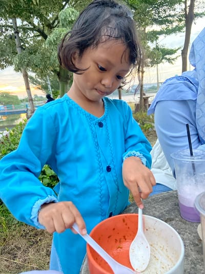 An outdoor scene in Kalitengah, Indonesia, depicts a young child in the foreground interacting with food. The child, with dark hair, wears a long-sleeved bright blue top with dark blue buttons and zigzag stitching, and matching blue pants. She is positioned facing downward, holding a white plastic spoon over an orange bowl containing remnants of food. A second white plastic spoon is present in an adjacent white bowl, also containing food residue. To the child's right, the partial figure of an adult is visible, dressed in a light blue top and patterned hijab. A transparent plastic cup containing a purple beverage and a black straw rests on a gray surface next to the adult. The background features a body of water with a light blue and white bridge, bordered by trees with green and light foliage. Another distant figure is partially visible near the bridge. The sky exhibits orange and yellow hues, indicating either sunrise or sunset. The ground consists of grass or dirt.