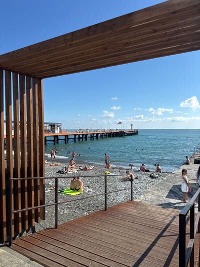 A wide shot captures a crowded beach and pier on a sunny day in Adlerskiy, Russia. The foreground features a wooden pergola, its planks casting shadows on a wooden walkway and a pebbled beach. Numerous people are scattered across the beach, some sunbathing on towels and mats, others wading in the turquoise water. A long, orange pier extends into the sea, lined with people and leading to a building at its end, where a red lighthouse is visible. The sky is a bright blue, dotted with a few white clouds. The overall scene depicts a lively and popular beach destination.
