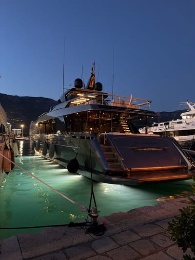The image depicts a large, luxurious yacht named "RIVIERA" docked at a marina in Budva, Montenegro, during twilight. The yacht is sleek and modern, with dark grey hull and multiple decks illuminated by warm interior lights. The water around the yacht is a striking, vibrant turquoise, lit by underwater lights that cast an ethereal glow. Several large, grey fenders are attached to the yacht's hull, protecting it from the dock. A thick rope is secured to a bollard on the stone quay, leading to the yacht. In the background, the lights of Budva's waterfront city can be seen along the base of a mountain range. Another vessel, a white passenger ferry with "KOMPAS" visible on its side, is docked nearby. The sky is a deep, clear blue, indicative of the early evening. No people are visible in the scene.