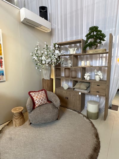 The image displays an interior setting, likely a living space or showroom, rotated 90 degrees clockwise. A large, rustic wooden shelving unit, featuring both open shelves and closed cabinets/drawers, dominates the central part of the frame. It holds various decorative items, including a small bonsai tree, woven baskets, cream and white ceramic sculptures resembling coral, brown cylindrical objects, and a patterned cushion.

To the right of the shelving unit, a soft, textured grey armchair or bean bag is placed on a light-colored, shaggy round rug. A prominent accent pillow with an orange and white geometric pattern and a reddish-brown fringed edge rests on the armchair. A small, light-colored cylindrical stool or side table is visible behind the armchair, and a wooden pedestal-like object sits on the rug.

On the floor to the left, a tall vase filled with white artificial flowers or branches is positioned in front of the shelving unit. Mounted on the wall above, a white air conditioning unit is visible next to a black speaker or similar device. The background features soft, translucent white curtains or blinds, indicating natural light. The overall color palette is neutral, dominated by shades of beige, brown, grey, and white. Some blurry text, possibly "U2" and "HO[...]", is visible on books or magazines on a lower surface in the bottom left corner. No people are present, and the scene conveys a calm, styled atmosphere.