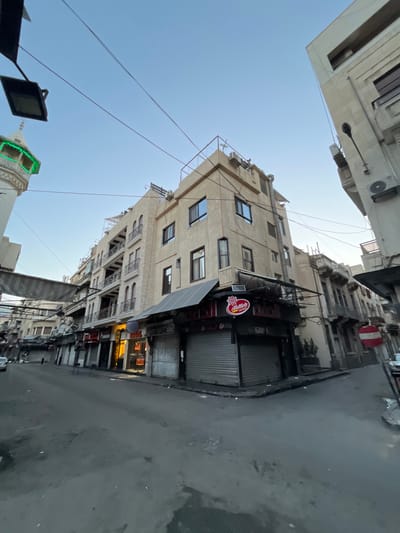 The image presents a narrow urban street scene in Al-Muhajerin Municipality, Syria, captured from a low-angle perspective looking upwards. Multi-story buildings with light-colored, possibly stuccoed, facades line both sides of the street. These structures feature numerous windows and some have small, ornate balconies. Attached to the building walls are several outdoor air conditioning units.
At ground level, multiple shopfronts are visible, mostly secured by metal roller shutters, indicating they are closed. A prominent circular red sign hangs from one building, featuring a white logo resembling a ship's wheel. Text on this sign includes "Alateh" (or "Al A'teh") and "Al-A'teh Food" (or "Fod").
The sky overhead is clear and bright blue, suggesting daytime. Numerous utility cables crisscross the expanse between the buildings. In the lower-left quadrant of the image, part of a mosque minaret is visible, distinguished by glowing green decorative lighting. The street itself is paved with asphalt or concrete, appearing somewhat worn and showing various marks or slight dampness. There are no people visible, and no overt activity is taking place, creating a quiet atmosphere.
