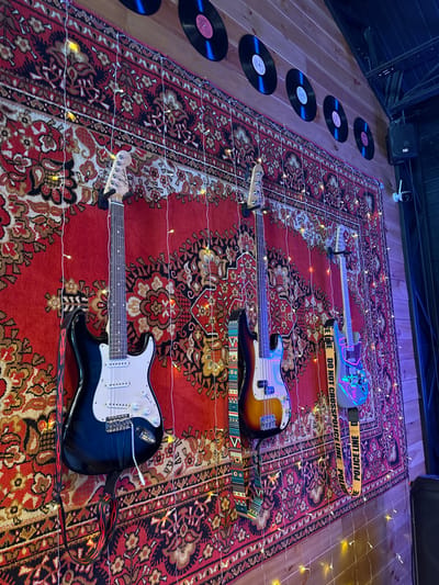 In Manzherok, Russia, a wall is adorned with a large, red Persian rug featuring intricate floral patterns. Several electric guitars are mounted on the rug. To the left, a black and white electric guitar is prominently displayed. In the center, a bass guitar with a sunburst finish is visible. To the right, a guitar with a unique, colorful neon-like finish is mounted. Above the guitars, on the wooden planked wall, several black vinyl records are hung. The scene is further decorated with strands of fairy lights that cast a warm glow. A yellow "POLICE LINE DO NOT CROSS" tape is draped across one of the guitars. The environment appears to be indoors, possibly a music studio or a themed room.
