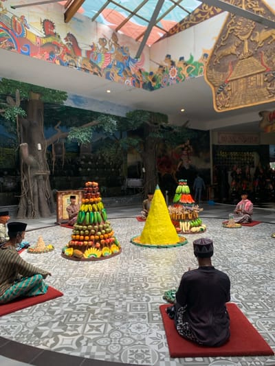 The image shows a circular room with a tiled floor. There are several men sitting around a circle with their legs crossed, holding offerings of fruits and vegetables in front of them. Four mounds of various fruits, vegetables, and other offerings are arranged in the circle. Two of the offerings are large conical shapes made of yellow paste. There are large murals on the wall depicting scenes of people riding horses. The ceiling is covered in a glass skylight. The room is located at the Museum Angkut, a transportation museum in Batu, Indonesia, at latitude -7.884 and longitude 112.525.