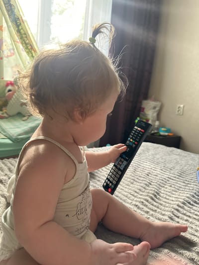 The image shows a toddler, seen from the side, sitting on a bed and holding a television remote control. The toddler has blond hair pulled into a ponytail and is wearing a light-colored tank top. The bed is covered with a gray, textured comforter. In the background, sunlight streams through a window with sheer curtains, illuminating the room. A dark curtain hangs to the right of the window. A bedside table with some items on it is visible in the distance, and a light switch is mounted on the wall. The overall scene appears to be in a domestic setting, likely a bedroom. The image does not contain any specific visual elements that would allow for an accurate determination of its location within the city of Sayanogorsk, Russia.