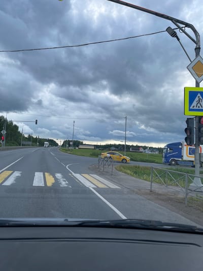 The image, apparently taken from inside a vehicle, shows a view of a road in Novyi Byt, Russia. The road is asphalt and has white and yellow lines marking the lanes. A pedestrian crossing with white and yellow stripes is visible. A yellow taxi cab is parked on the side of the road. Traffic lights and road signs, including a pedestrian crossing sign and a diamond-shaped sign, are present. A blue truck with the EU flag on it is also parked on the side of the road. The sky is overcast with heavy clouds. Trees and other vegetation line the road. The image is rotated counter-clockwise.