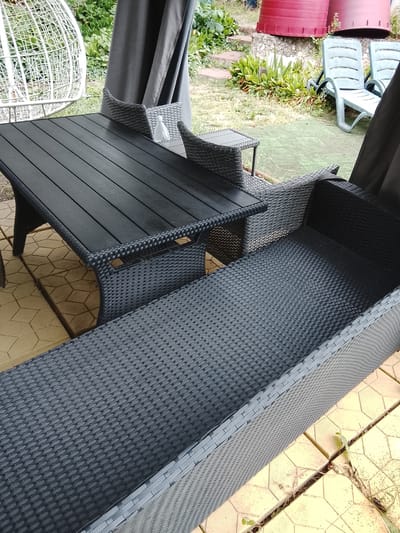 The image depicts an outdoor seating area on a paved patio, likely in the city of Codru, Moldova. In the foreground, a dark gray wicker-style bench or sofa is prominently featured, with its textured seating surface filling much of the frame. Behind the bench, a dark gray wicker-style dining table with a slatted top is visible. To the left of the table, part of a white, egg-shaped hanging chair is in the frame. In the background, the scene opens up to a yard with green foliage, a grey canopy or curtains, and two light blue lounge chairs. Two large, red cylindrical objects, possibly planters or barrels, are situated on a stone wall in the distance. The overall impression is of a relaxed outdoor living space, possibly a backyard or patio area. There are no people visible in the image.